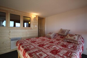 A bedroom with a wooden wall, a window, a bed with patterned bedding, and a wall-mounted heater.