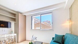 Una sala moderna con sofá, televisor montado en la pared, comedor con sillas y ventana con vistas a edificios.