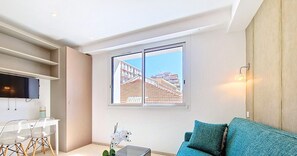 Een moderne woonkamer met een bank, een aan de muur bevestigde televisie, een eethoek met stoelen en een raam met uitzicht op gebouwen.