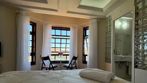 Una cama grande con cubrecama acolchado, dos sillas, una mesa con un florero y vistas al exterior a través de amplias ventanas con cortinas.