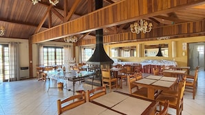 Un comedor espacioso con altos techos de madera y múltiples candelabros. Las mesas están dispuestas en filas con sillas, y se observa una chimenea grande en el centro.