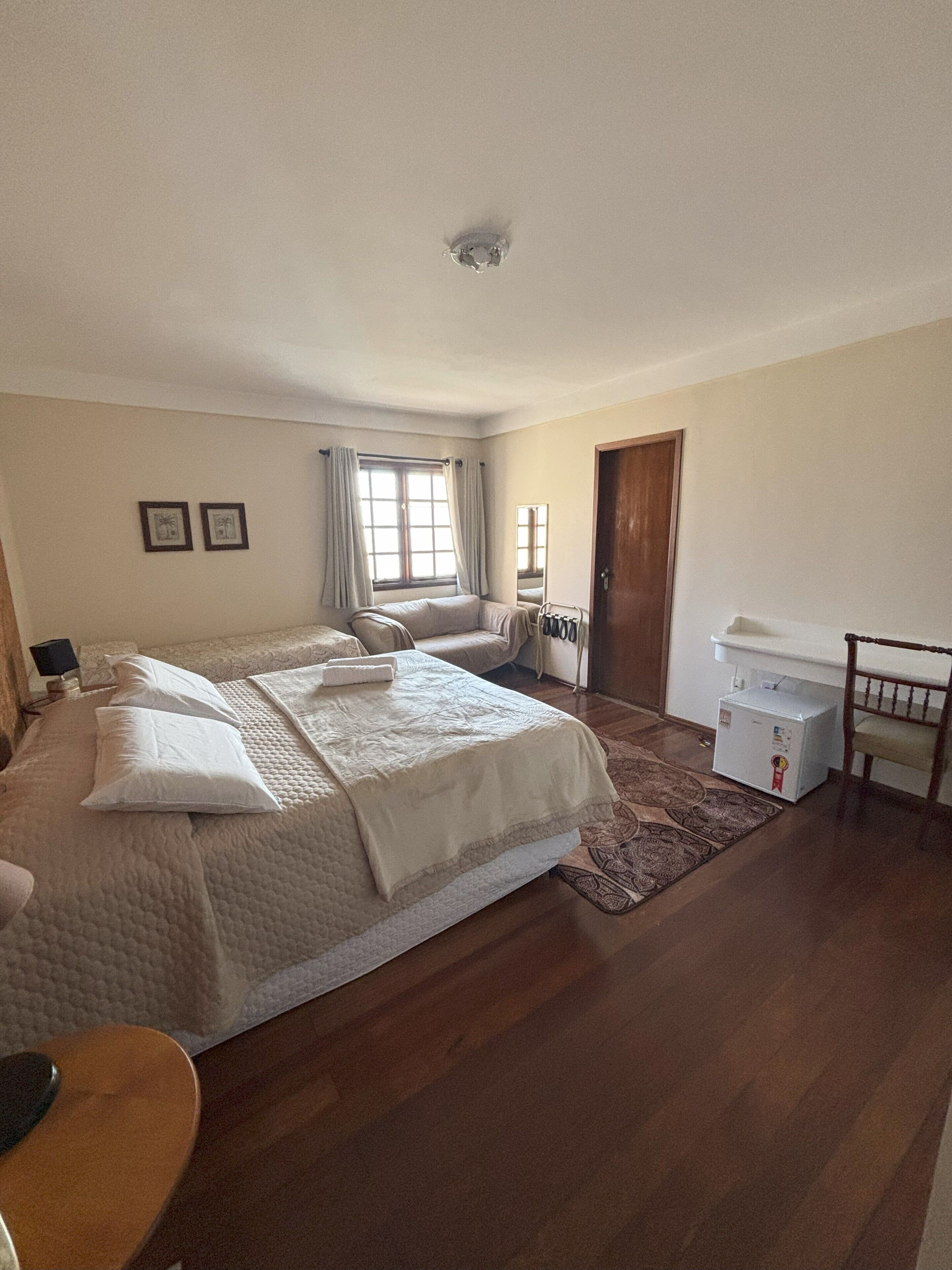 Een slaapkamer met een groot bed, een klein tafeltje, een stoel, een raam met gordijnen, een deur en een witte koelkast.