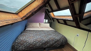 Ein Schlafzimmer mit schrägen Wänden, einem Bett mit gemusterten Textilien und einem Fenster mit Blick zum Himmel.