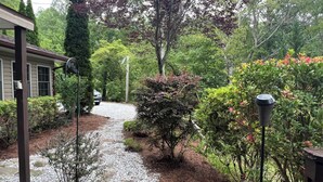 Un chemin de gravier mène à une maison dotée d’un porche abrité, entourée d’une végétation luxuriante et de diverses plantes.