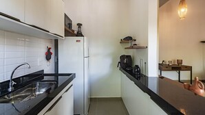Μια μικρή κουζίνα με λευκά ντουλάπια, έναν μαύρο πάγκο και ένα ψυγείο. Πάνω από τον πάγκο υπάρχουν ράφια όπου είναι τοποθετημένη μια ηλεκτρική βραστήρα και μια καφετιέρα.
