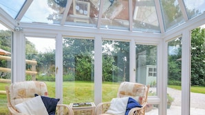 A conservatory with wicker furniture and glass doors, offering a view of a garden and a house. - Upper Court Cottage (Bude)