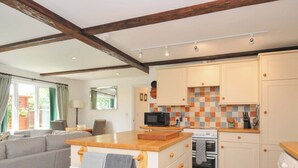 A cozy living room with a sofa, a wooden coffee table, and a radiator. The kitchen features a tiled backsplash, wooden cabinets, a microwave, and a washing machine. - Upper Court Cottage (Bude)