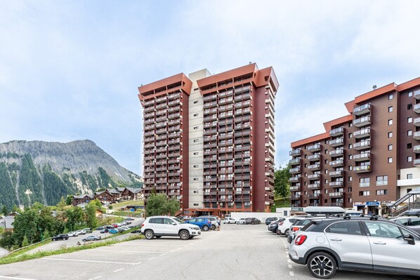 A multi-story hotel with balconies, a parking lot, and a mountainous backdrop.