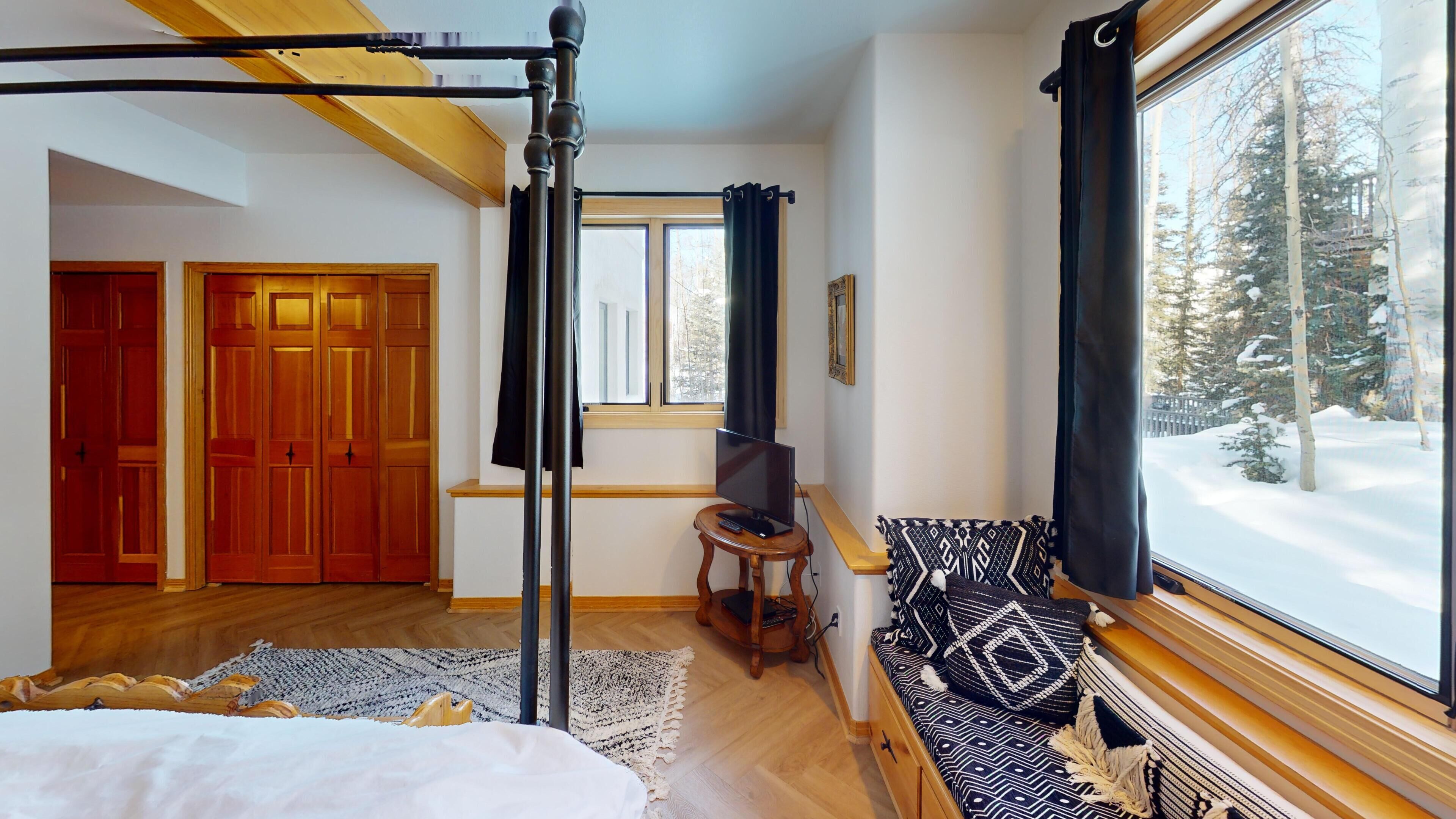 Une chambre à coucher avec un lit à baldaquin, un téléviseur posé sur une petite table, une fenêtre avec des rideaux et une vue sur une forêt enneigée.