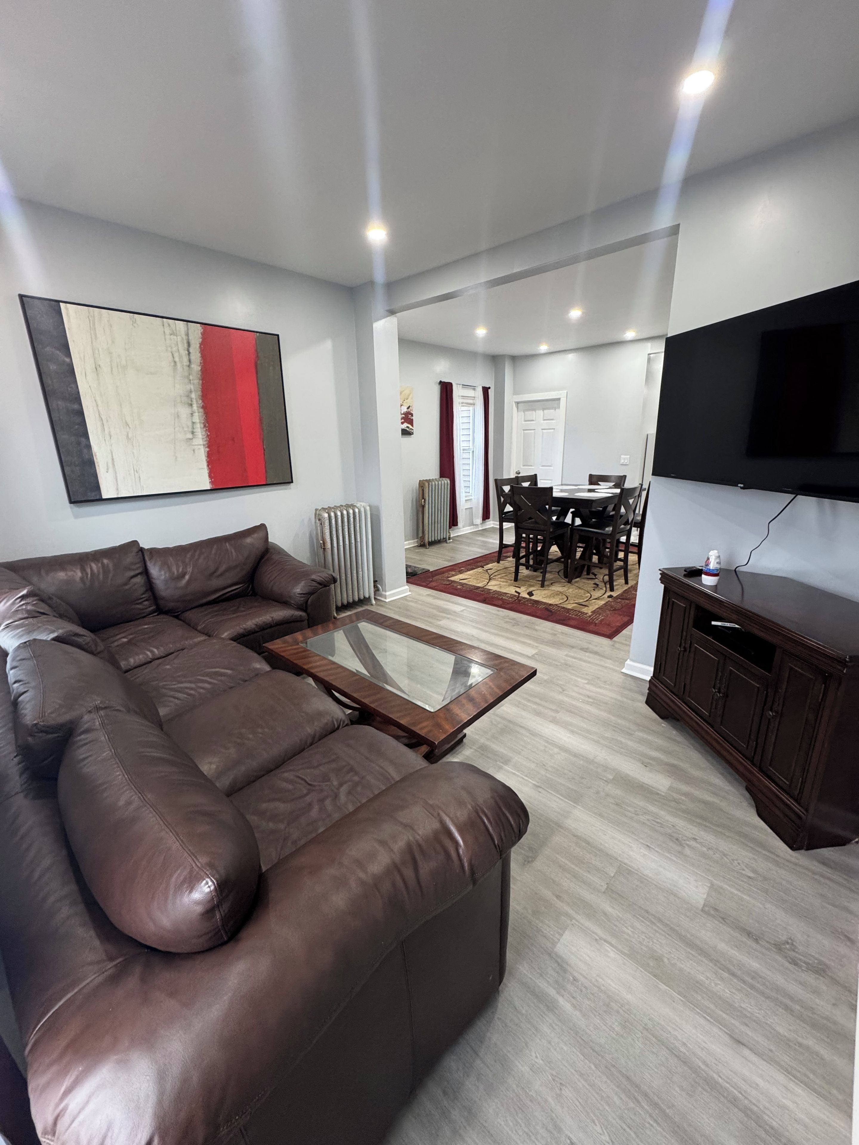 A modern living room with a leather sofa, a glass coffee table, a dining area with a table and chairs, and a flat-screen TV mounted on the wall.