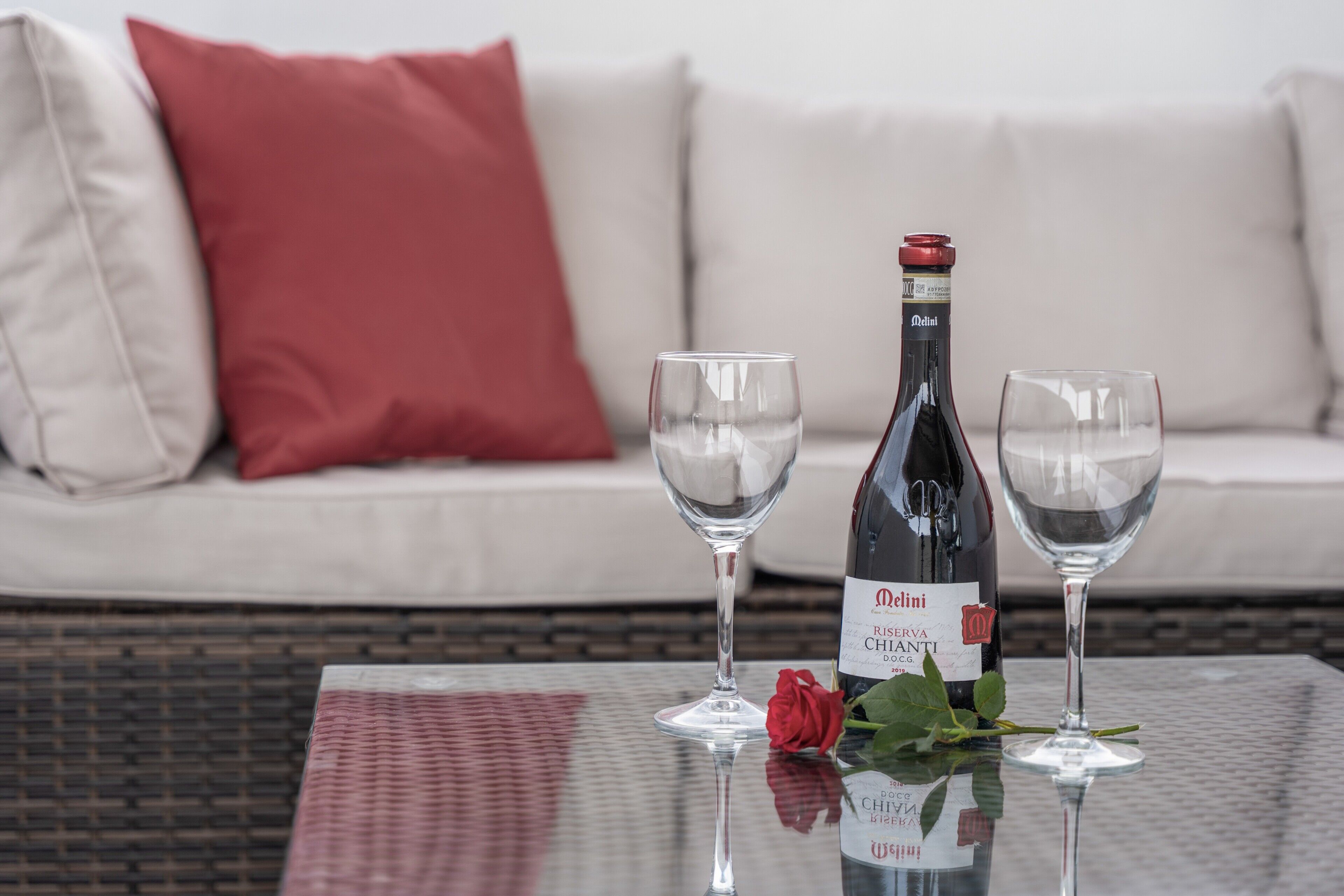 Une bouteille de vin, deux verres et une rose sur une table en verre, avec une chaise en osier et un canapé en arrière-plan.