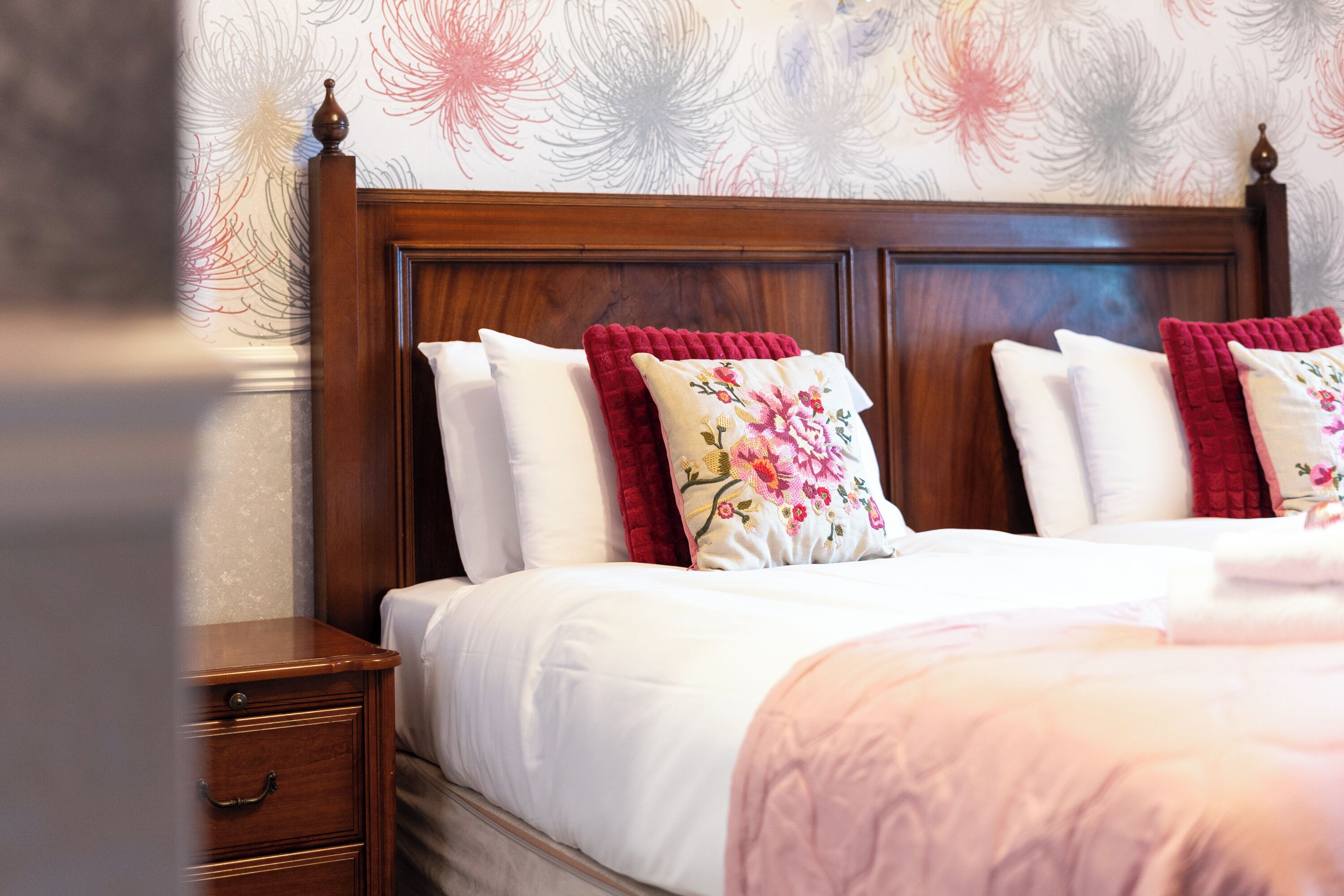 A neatly made bed with white and red pillows, a wooden headboard, and a floral wallpaper pattern.