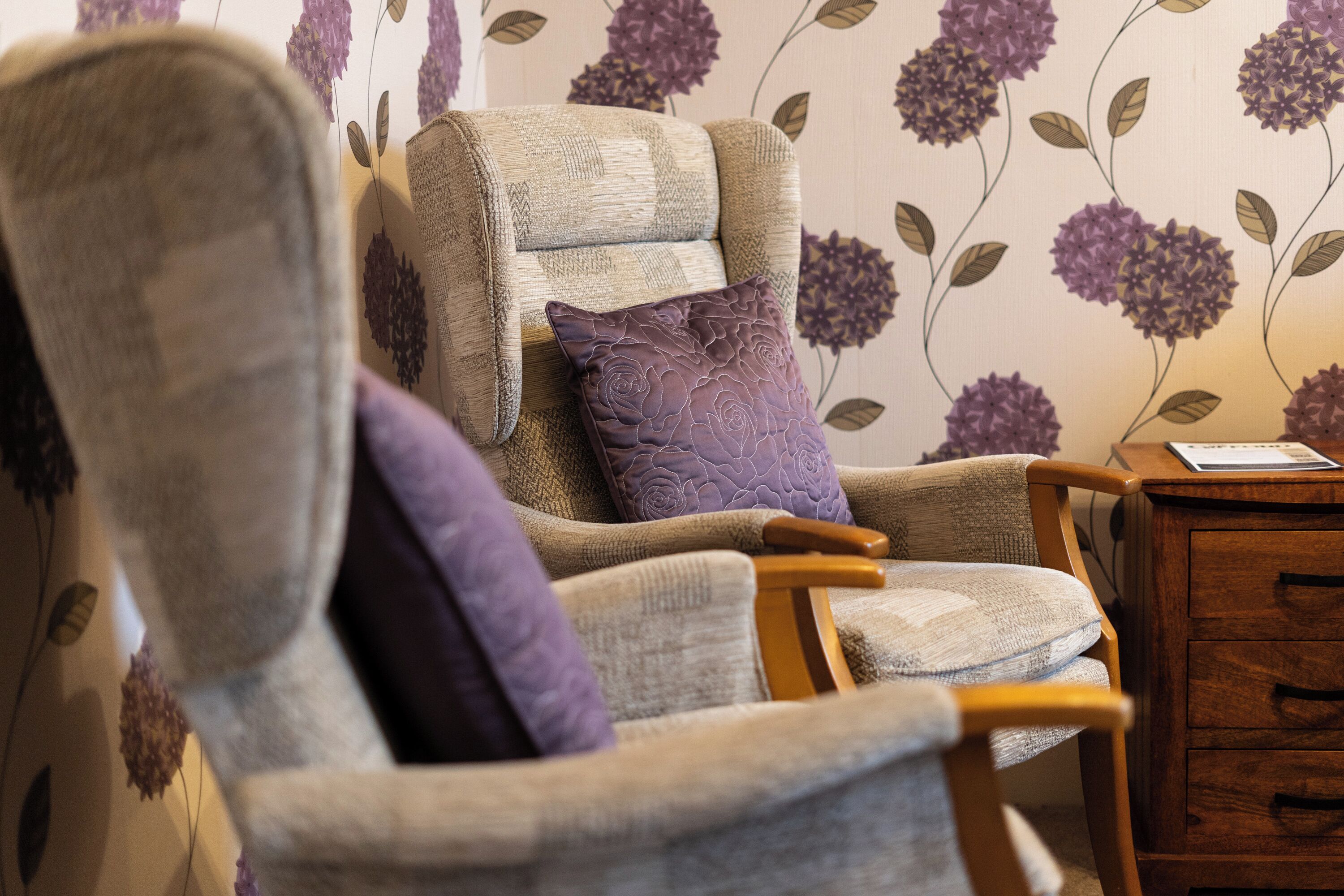 A chair with a cushion, a wooden side table, and a wall with floral wallpaper.