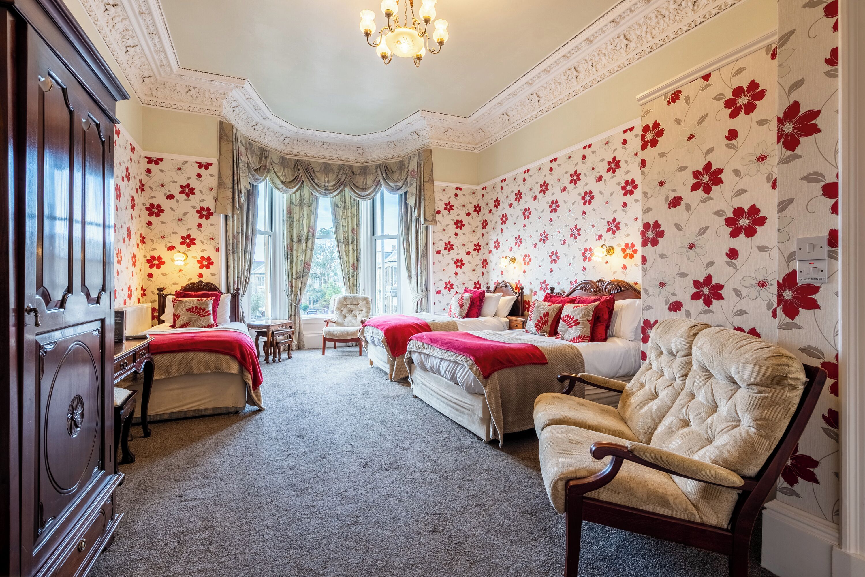A bedroom with floral wallpaper, a chandelier, a large bed, two armchairs, and a wooden dresser.