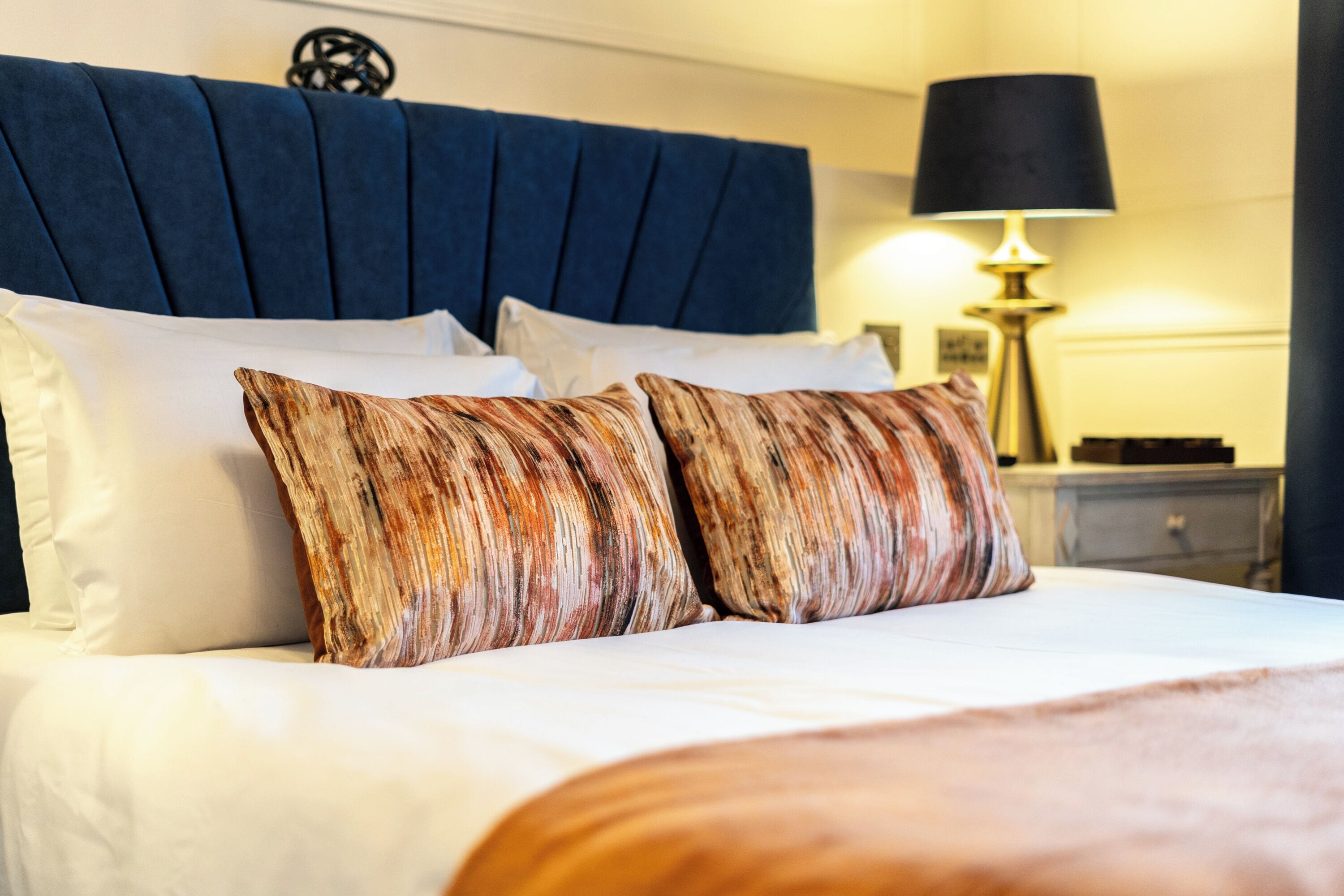 A neatly made bed with white linens and two patterned pillows, a dark blue upholstered headboard, and a bedside table with a lamp.