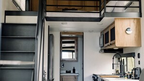 A compact, modern kitchen with a built-in microwave, a sink, and a stove. A staircase leads to an upper level with a wooden ceiling.