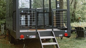 A black mobile home with a ramp and a small deck, surrounded by trees.