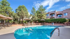 A swimming pool with lounge chairs and umbrellas, surrounded by trees and a building with balconies.