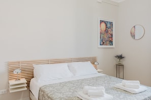 A neatly arranged bedroom with a bed, bedside tables, a bedside lamp, a framed artwork, a round mirror, and a small potted plant.