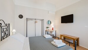 A hotel room with a bed, a desk, a chair, a TV, a clock, and a bench.
