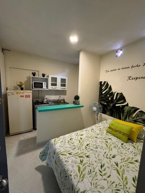 Habitación funcional con kitchenette, cama con colcha estampada de hojas y pared decorada con una planta grande y texto.