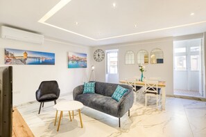 Un salon moderne avec un canapé gris, une table ronde blanche et un fauteuil noir. Deux tableaux ornent le mur, ainsi qu’une horloge et une fenêtre donnant sur la mer.