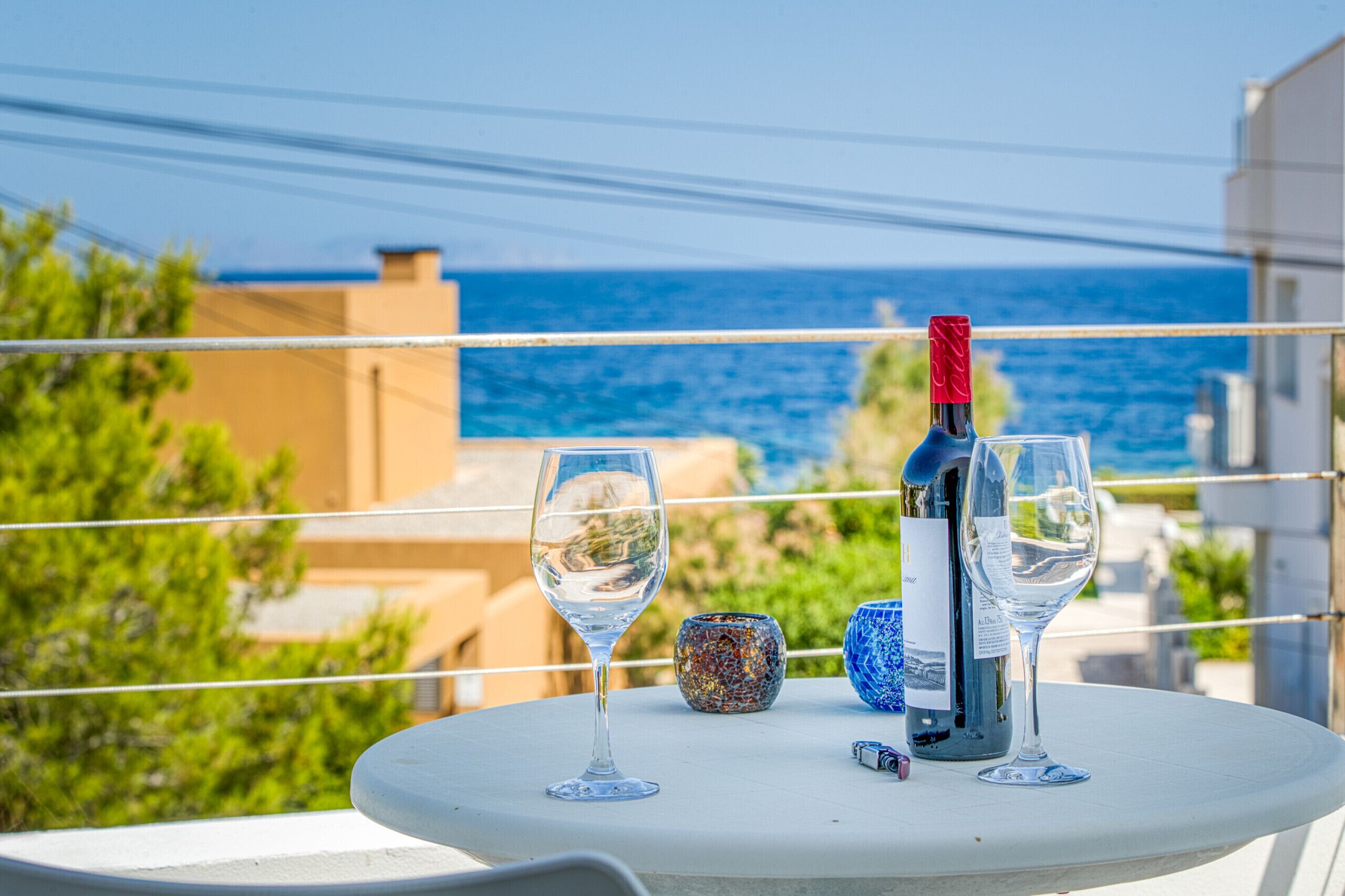A table with two wine glasses, a bottle, and a decorative item on a balcony overlooking the sea.