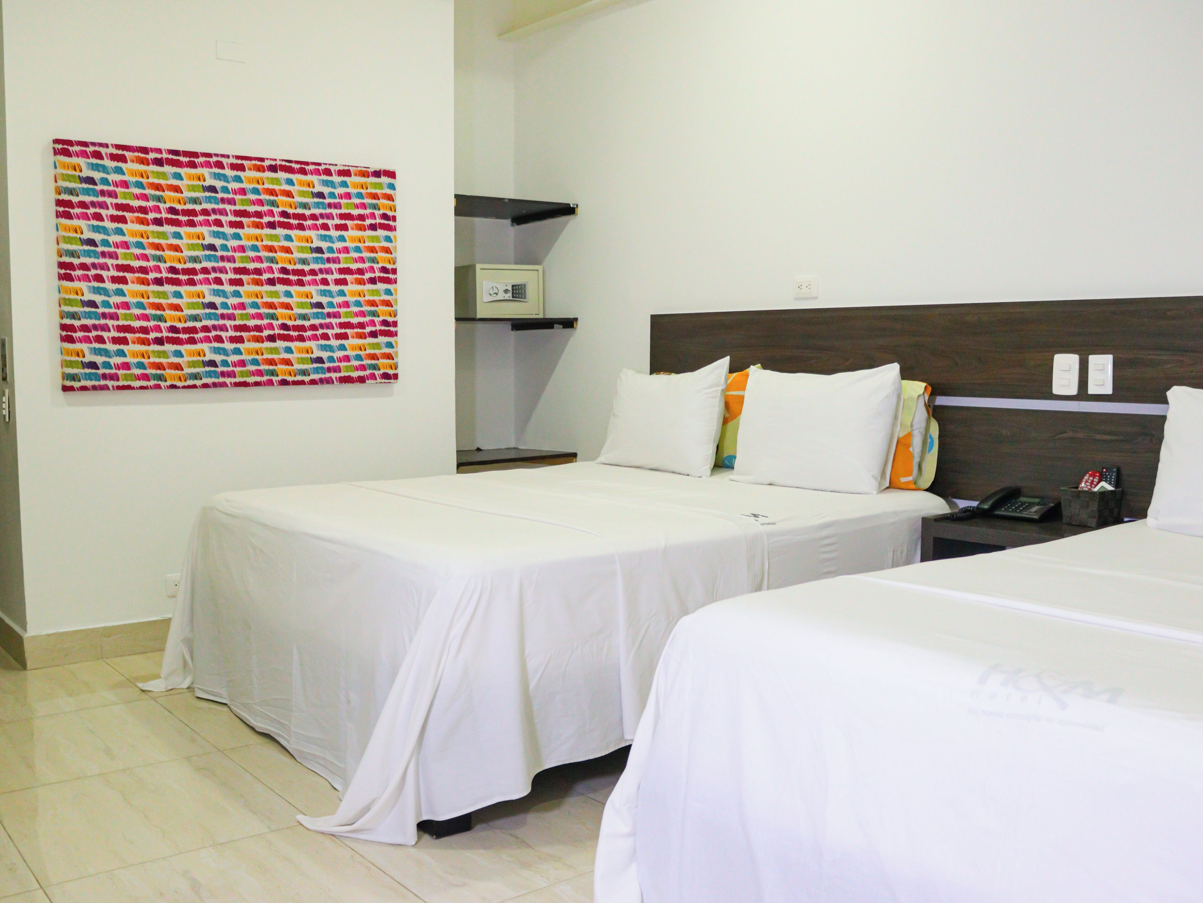A hotel room with two beds, white bedding, a colorful abstract painting on the wall, a small shelf with a phone and remote controls, and a plain white wall.