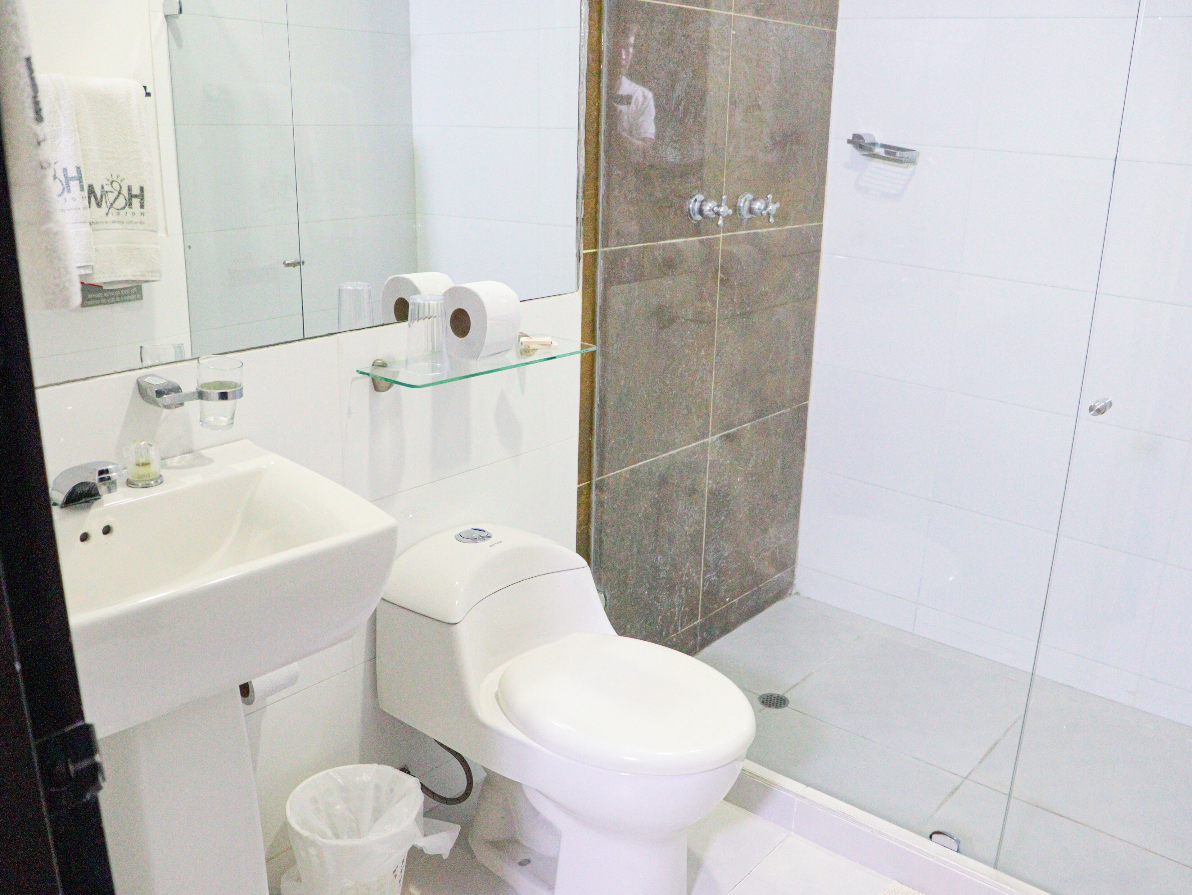 A bathroom with a white sink, toilet, and shower. There is a glass shelf above the toilet holding toilet paper and a glass of water.