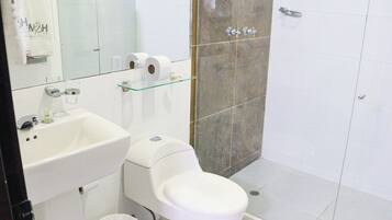 Un baño con lavamanos, inodoro y ducha blancos. Hay un estante de vidrio sobre el inodoro con papel higiénico y un vaso de agua.