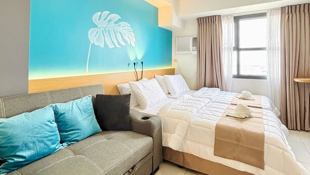 A hotel room with a bed, a sofa, a large wall mural, a window with curtains, and an air conditioning unit. - Spacious Studio @Horizon 101 with Aesthetics View (Cebu City)