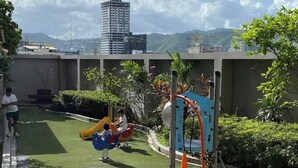 A rooftop playground with a seesaw and a swing set, surrounded by greenery and a view of a cityscape with a tall building. - Spacious Studio @Horizon 101 with Aesthetics View (Cebu City)