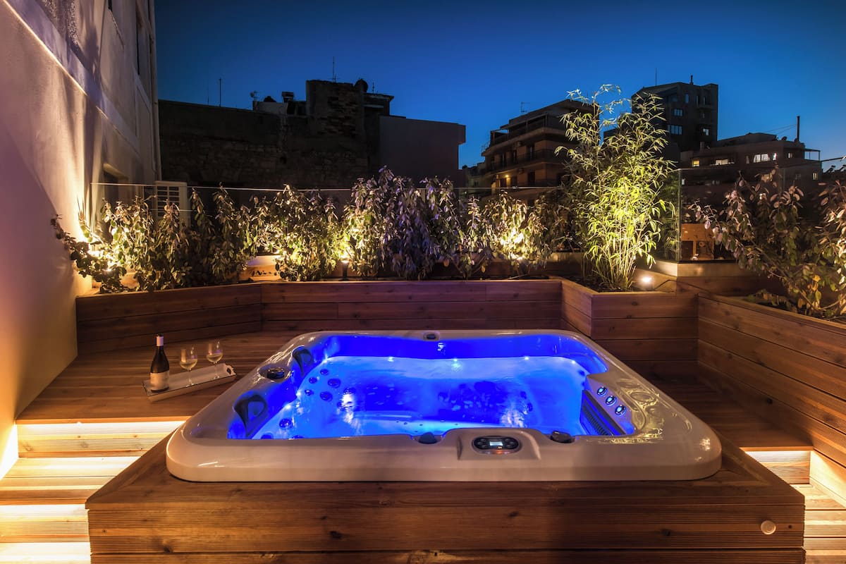 Un jacuzzi con muelle de madera y vista a edificios y plantas al anochecer.