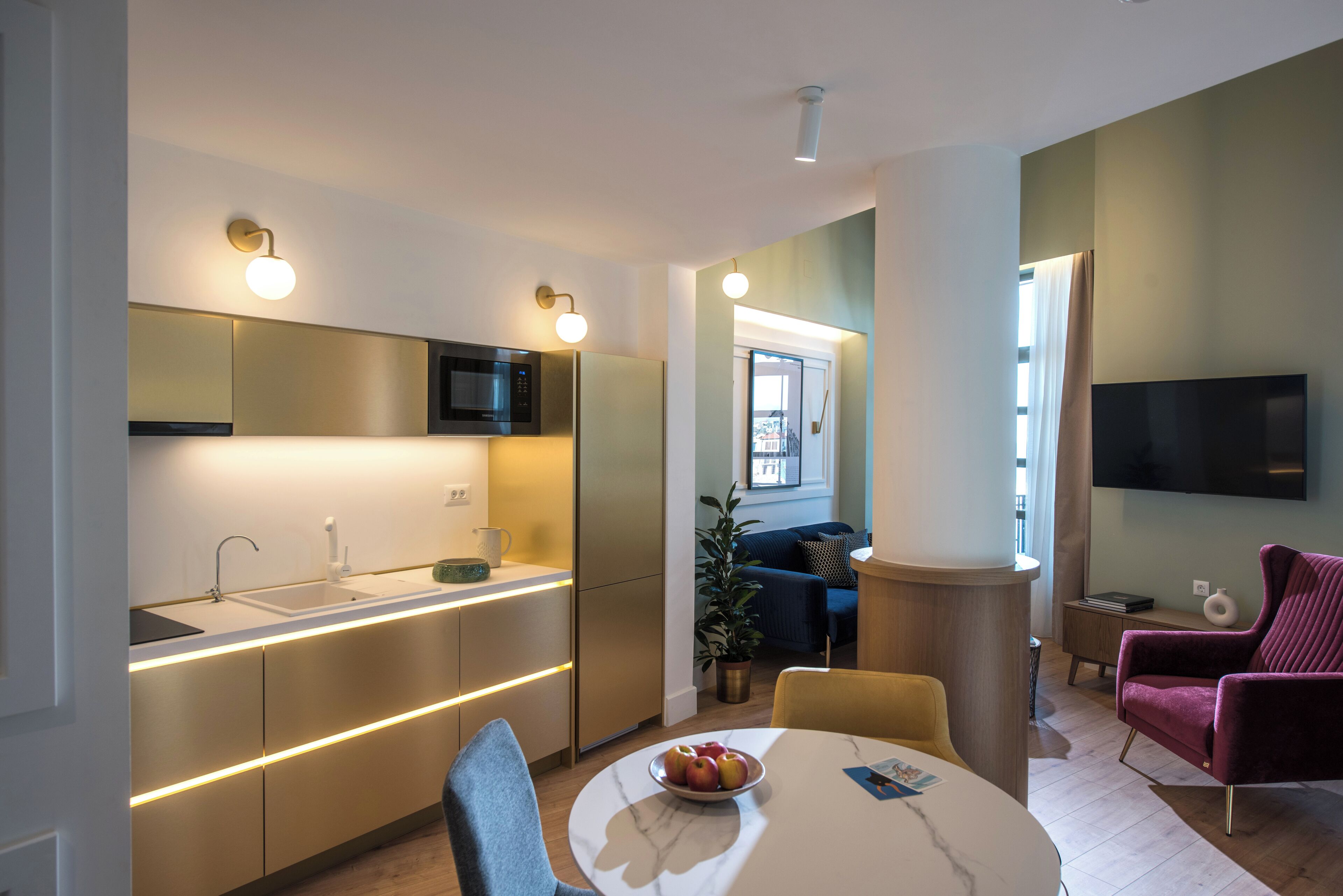 Una cocina moderna con electrodomésticos integrados, un comedor con una mesa redonda y sillas, y una sala de estar con sofá y televisor.