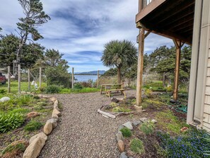 A gravel pathway leading to a wooden bench under a pergola, surrounded by a garden with various plants and a view of a lake.