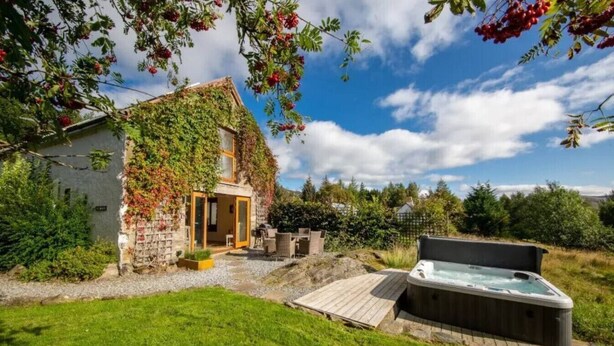 Un bâtiment rustique en pierre, doté d’un patio couvert et d’un jacuzzi, niché dans un jardin luxuriant.