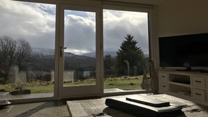 Ett rum med en stor skjutdörr i glas som leder ut till en vacker utomhusvy, en platt-TV på ett vitt TV-bord och ett litet bord med en bordsduk.