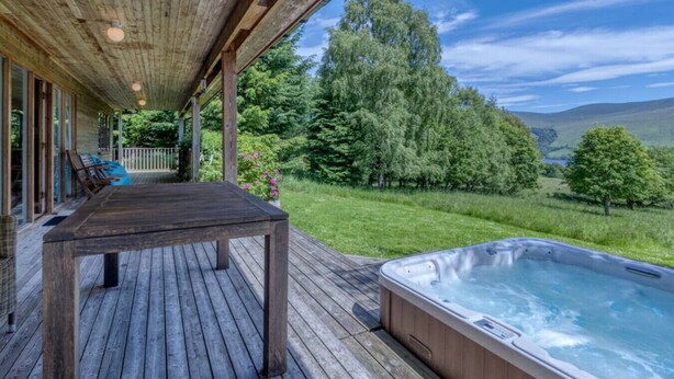 A wooden deck with a hot tub and a table, overlooking a lush green landscape and hills.