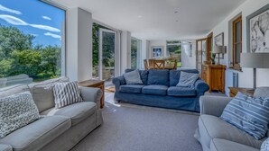 A spacious living room with two sofas, a coffee table, and a dining area with a table and chairs. Large windows provide a view of the outdoors.