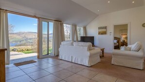A spacious living room with a large sliding glass door leading to a scenic outdoor view, a flat-screen TV, and a cozy seating area with two sofas. - Oak Cottage & Hot Tub, Loch Tay (Perth and Kinross)