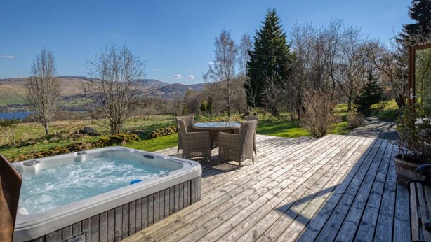 A wooden deck with a hot tub, wicker chairs, and a view of a lake and mountains. - Oak Cottage & Hot Tub, Loch Tay (Perth and Kinross)