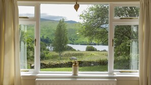 A room with a large window overlooking a scenic landscape with a lake, trees, and hills. - Oak Cottage & Hot Tub, Loch Tay (Perth and Kinross)