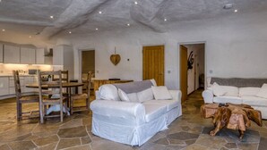 A spacious living area with a stone floor, white sofas, a wooden dining table, and a kitchen in the background.