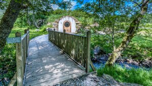 A wooden bridge leading to a rustic stone structure with a circular window, surrounded by lush greenery and a clear blue sky.
