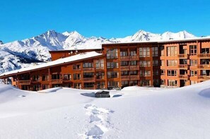 A multi-story hotel with a wooden facade, large windows, and balconies, set against a backdrop of snow-covered mountains.