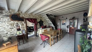 Un intérieur rustique avec un mur en pierre, des poutres en bois au plafond, un coin repas avec une table et des chaises en bois, une cuisine avec un comptoir et des appareils électroménagers, et un escalier menant à un autre niveau.
