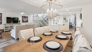 Una zona menjador moderna amb una taula de fusta preparada per a quatre persones, envoltada de cadires beix. Sobre la taula penja un penjoll únic, i es veu una escala al fons que condueix a un nivell superior.