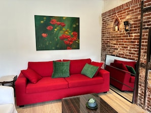 Ein rotes Sofa mit grünen Kissen, ein kleiner Tisch mit einer Pflanze und eine Ziegelwand mit einem Bild roter Blumen.