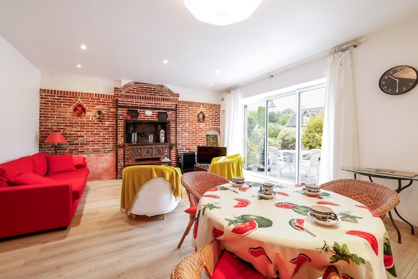 A living room with a red sofa, a dining table with a watermelon-themed tablecloth, a brick wall, and a view through a glass door.