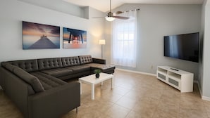 Un salon moderne avec un canapé d’angle, un téléviseur à écran plat, un ventilateur de plafond et des œuvres d’art accrochées au mur.